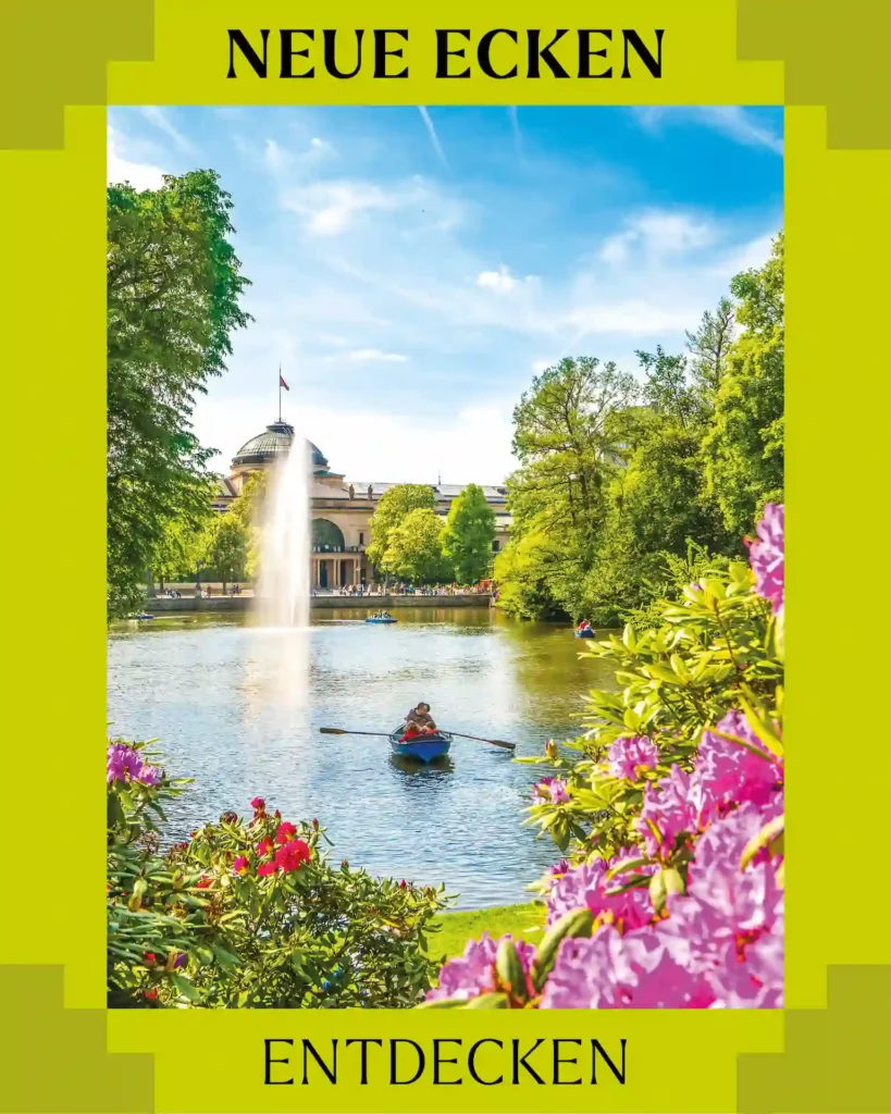 Hellblauer Himmel. ein See mit 2 Paddelbooten und Springbrunnen dahinter ein Gebäude. Vorne auf dem Bild sind Blumensträucher mit pinken und roten Blüten